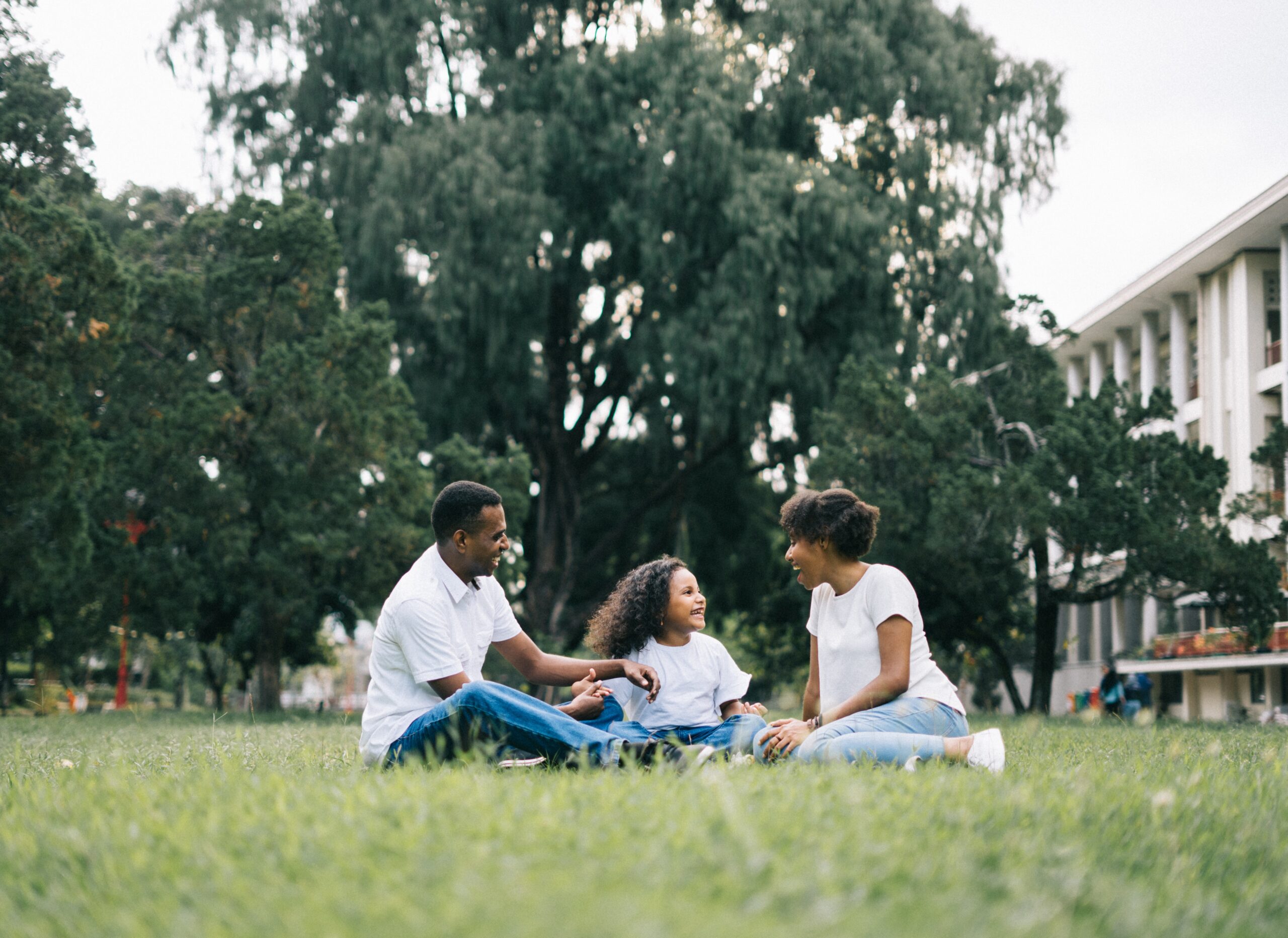 mother, father and daughter sitting in the grass smiling and looking at one another. 